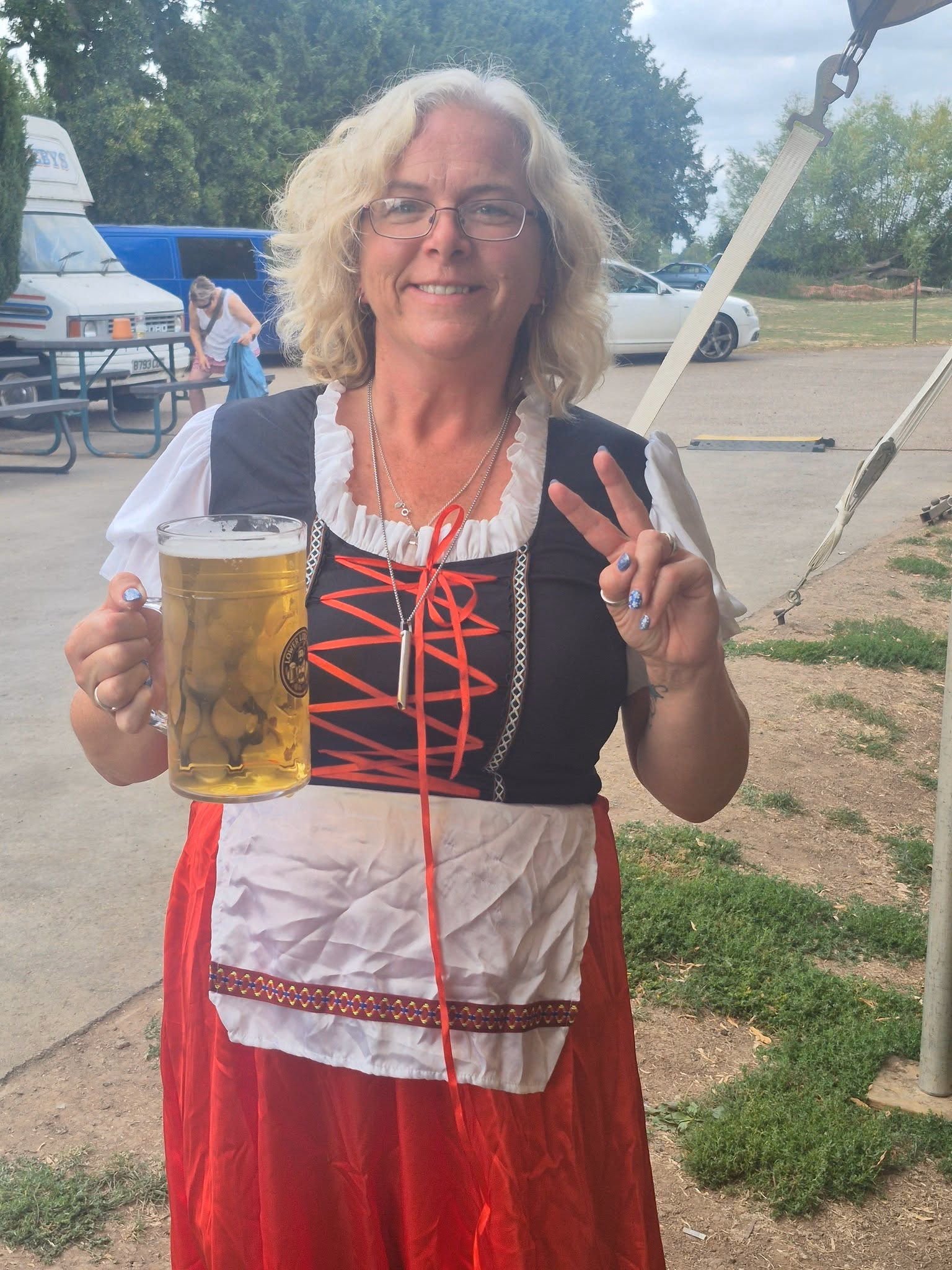 Festival visitor in red, white and black Bavarian-style dress holding a large glass of lager and making a peace sign.