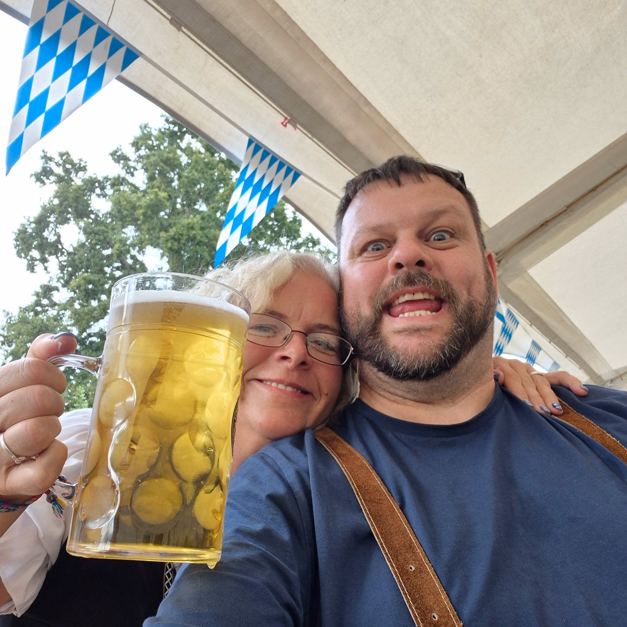Two Oktoberfest visitors smiling for a selfie beneath Bavarian bunting, one holding a large glass of lager.