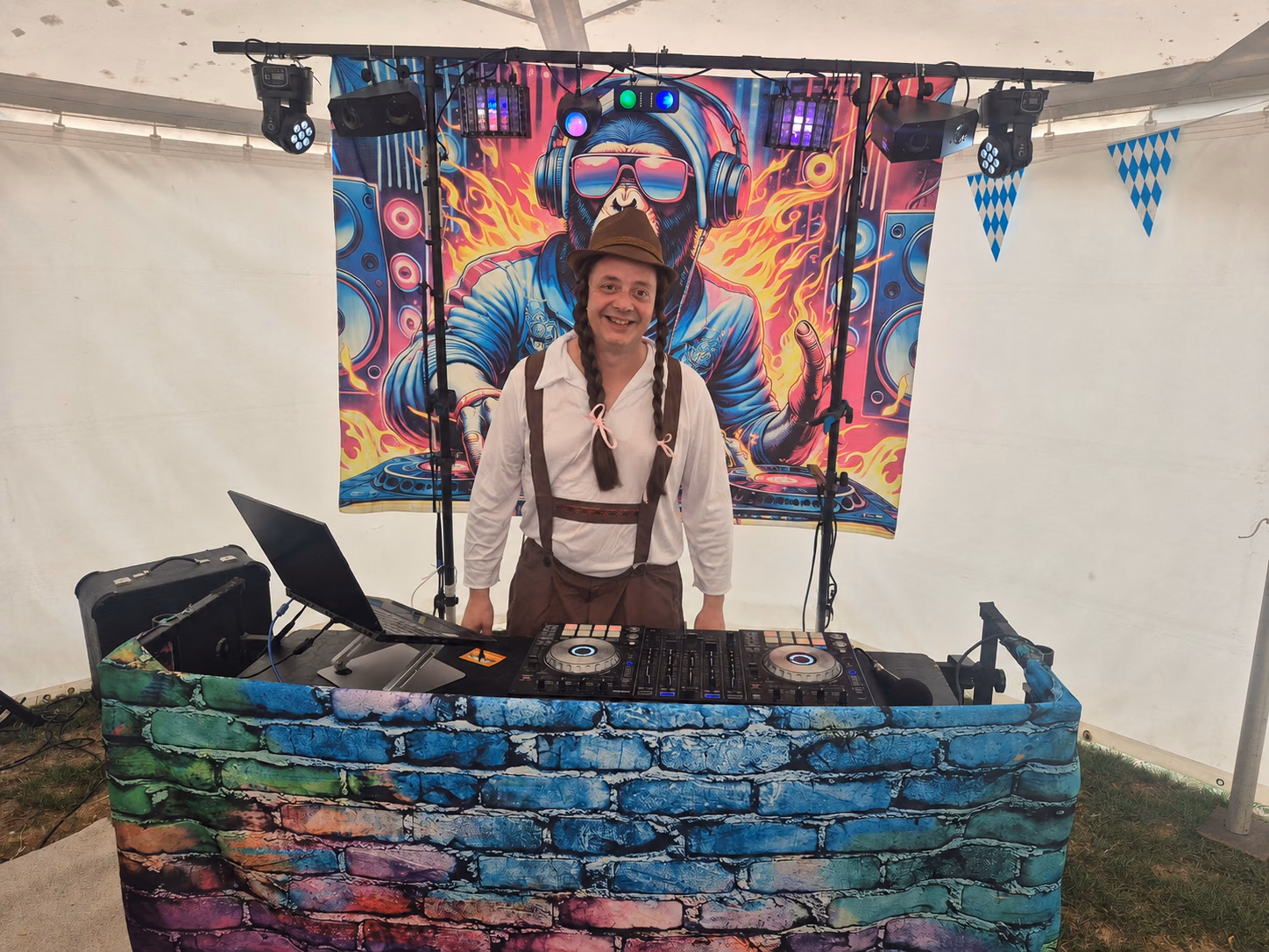 Festival DJ smiling behind a colourful DJ booth beneath blue-and-white Bavarian bunting.