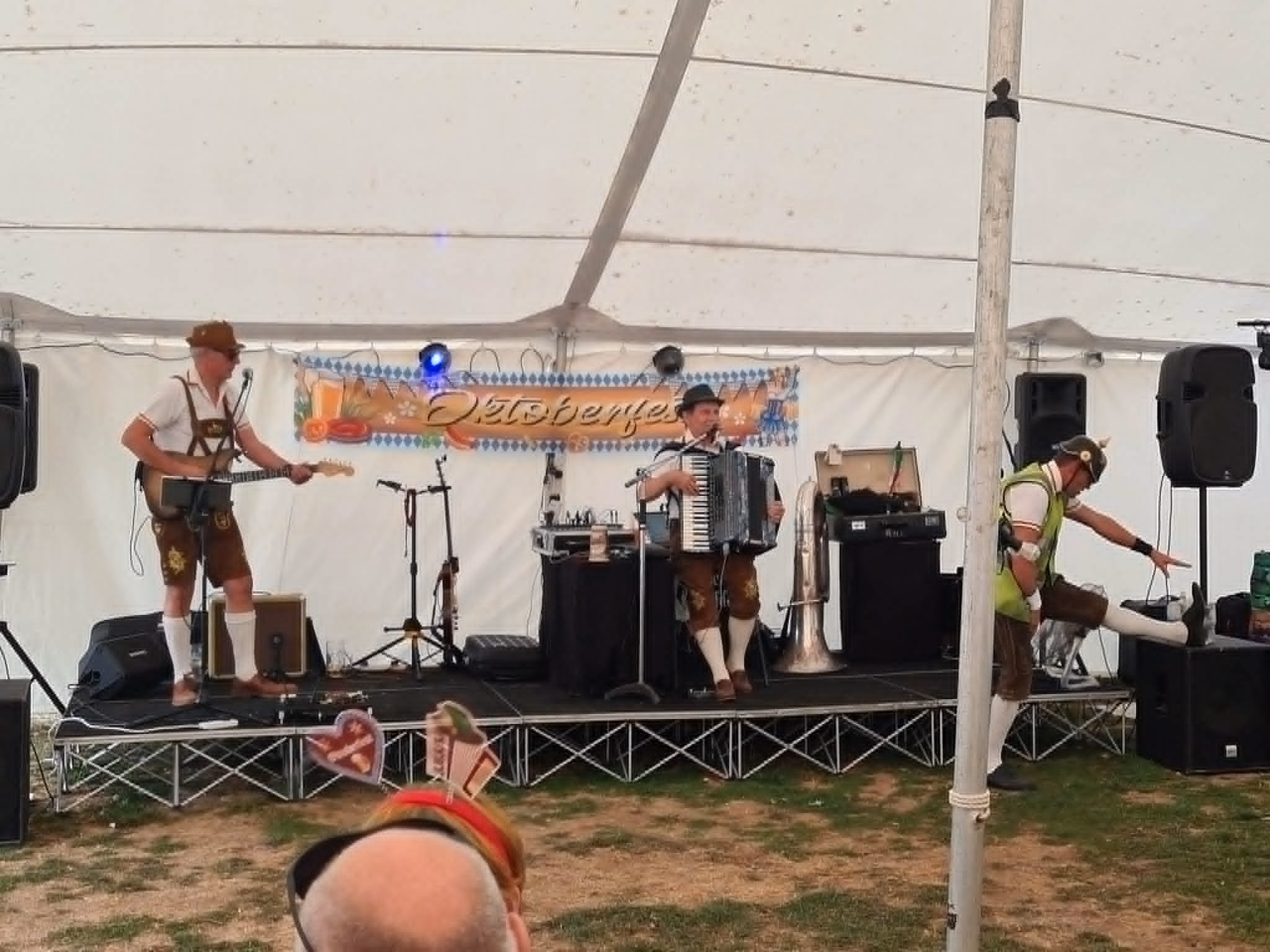 Three Oompah musicians performing with guitar and accordion on the marquee stage at the 2025 Lower Lode Inn Oktoberfest.
